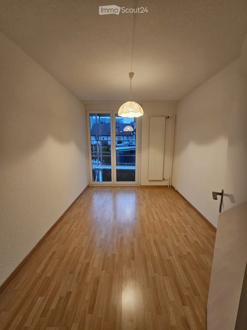 Empty room with wooden flooring, white walls, and a large window with a view of the outside. A single hanging light fixture is present.
