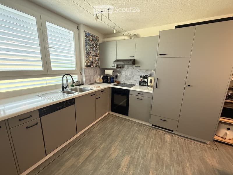 Modern kitchen with light grey cabinets, white countertops, stainless steel sink and faucet, built-in oven, dishwasher, and a window with blinds.