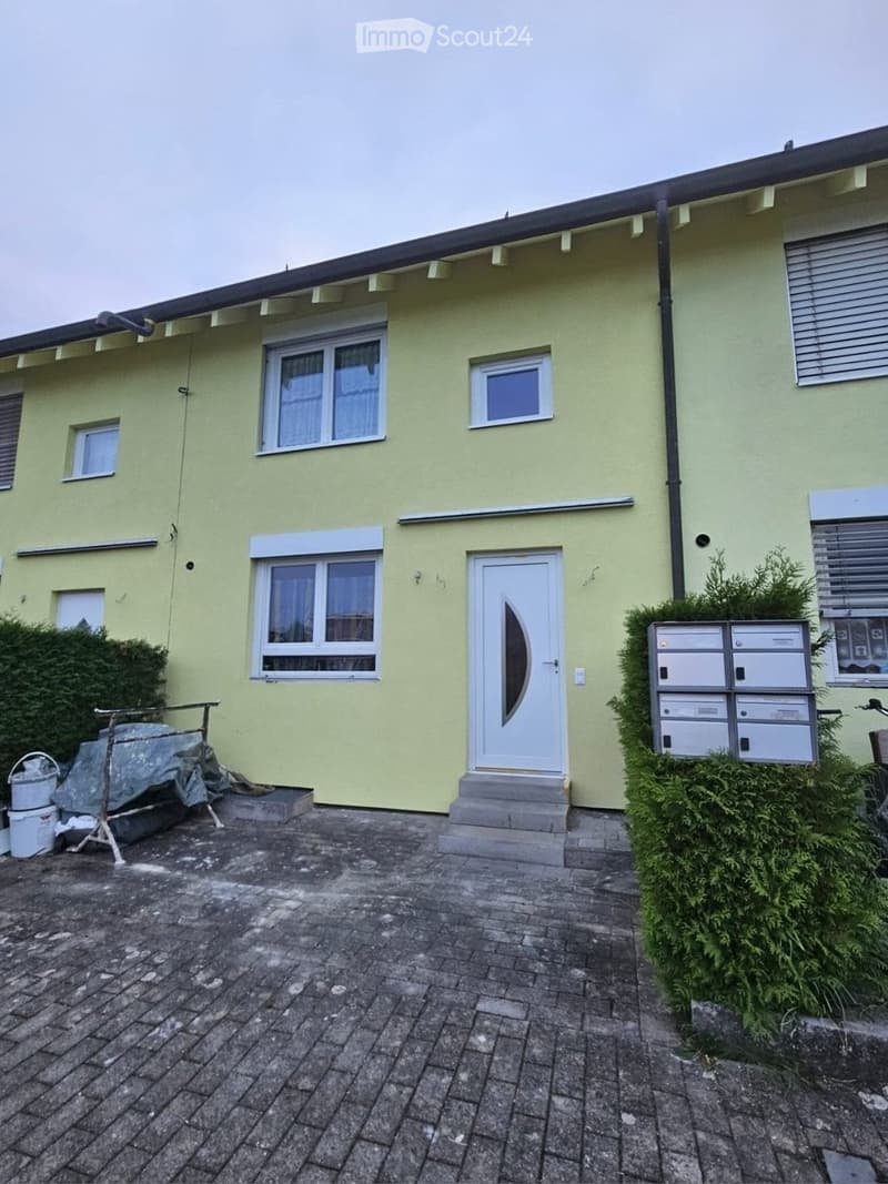 Two-story house with light green exterior, multiple windows, and a front door with a decorative glass panel. The entrance has a small concrete step, and there is a mailbox unit to the right. The ground is paved with bricks, and there are some bushes and a