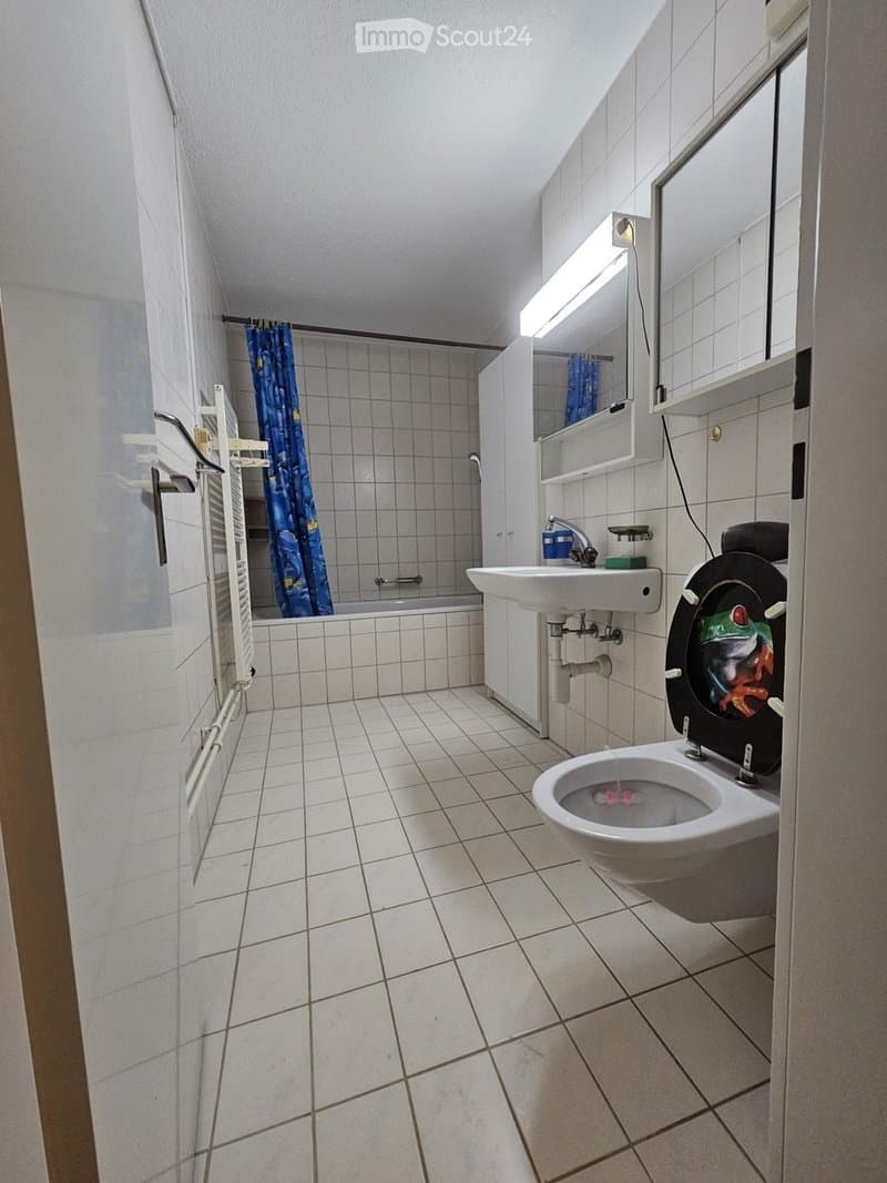 Bathroom with tiled walls and floor, featuring a toilet with a colorful seat cover, a sink with a mirror above, a bathtub with a blue curtain, and a radiator on the wall.