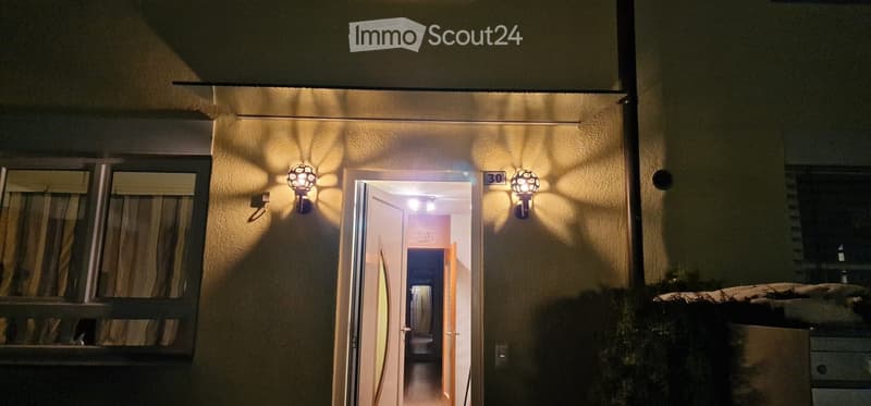 Nighttime view of a building entrance with illuminated wall-mounted lights, open doorway leading into a lit interior hallway.