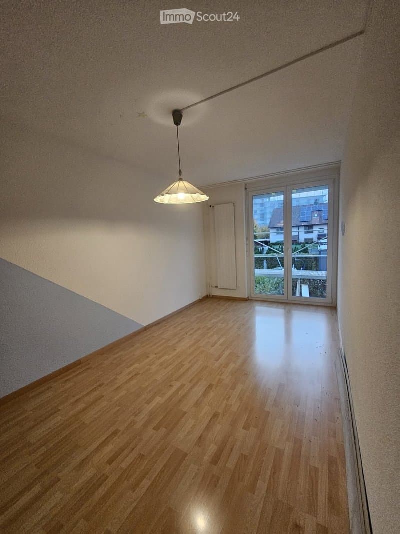 Empty room with wooden flooring, white walls, and a large window with a view of the outside. A single pendant light hangs from the ceiling.