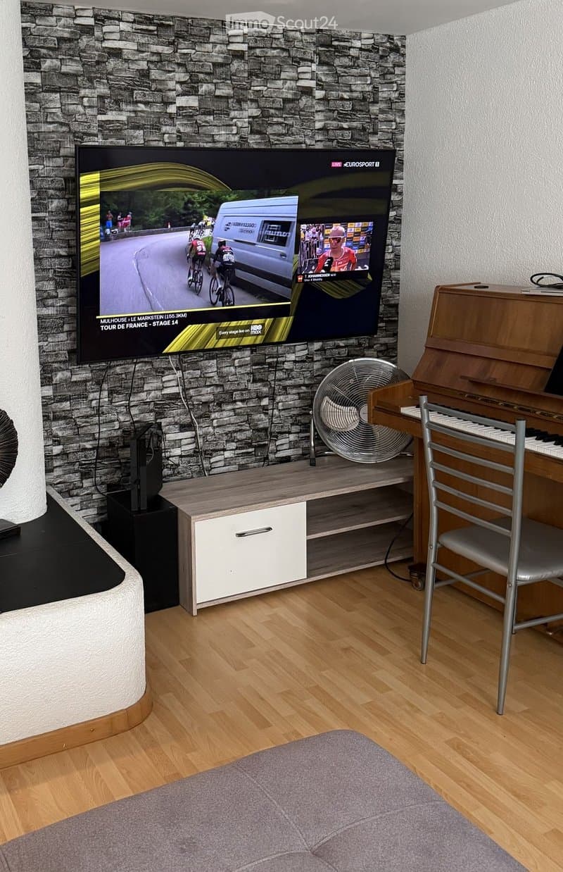 A modern living room with a large flat-screen TV mounted on a stone-textured wall, a wooden TV stand, a grey sofa, a small fan, and a piano with a chair.