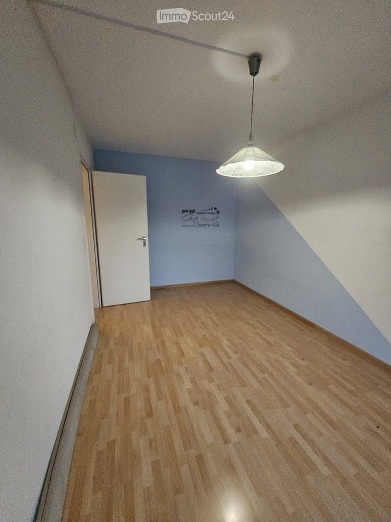 Empty room with wooden flooring, white walls, and a single hanging light fixture. The room has a slanted ceiling and a door leading to another space.