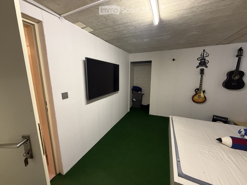 A small bedroom with a single bed, a mounted TV, and two guitars on the wall. The room has green carpeting and white walls.
