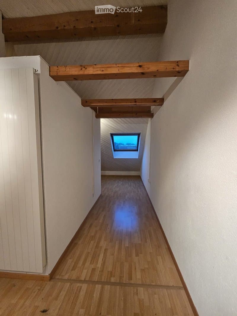A narrow hallway with wooden flooring and exposed wooden beams on the ceiling. A small skylight window at the end provides natural light.
