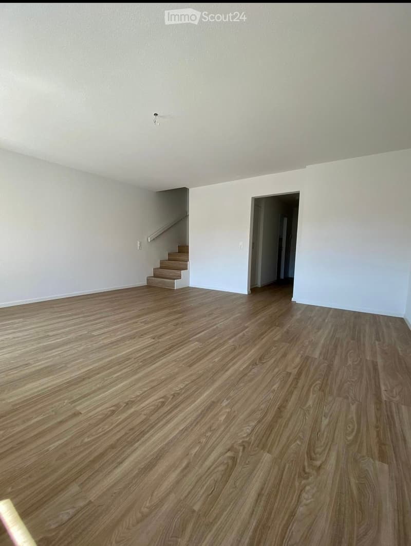 Empty room with light-colored walls and polished wooden flooring, featuring a staircase leading upwards and an open doorway to another area.