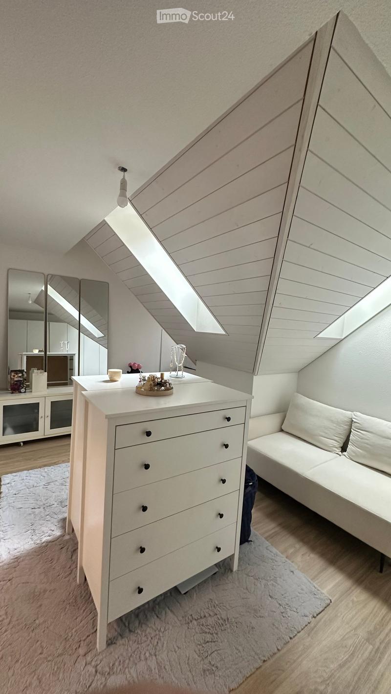 A modern, well-lit attic room with wooden slanted ceilings and large windows. Features a white dresser with multiple drawers, a small bench, a large mirror, and a decorative tray on the dresser.