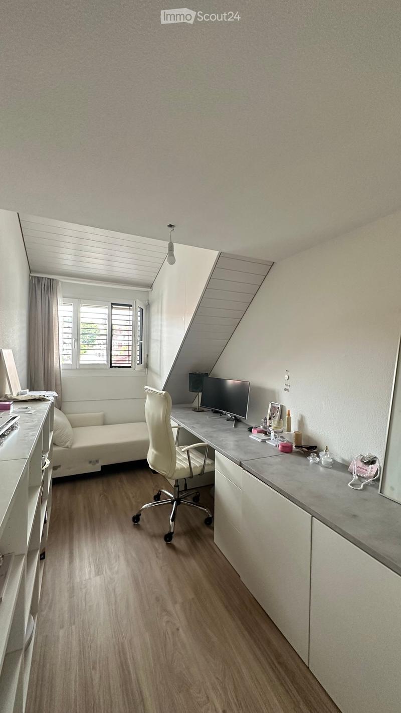 A modern, well-lit room with a slanted ceiling, featuring a white leather chair, a small sofa, a desk with a screen, and a lamp. The room has wooden flooring and large windows with blinds.