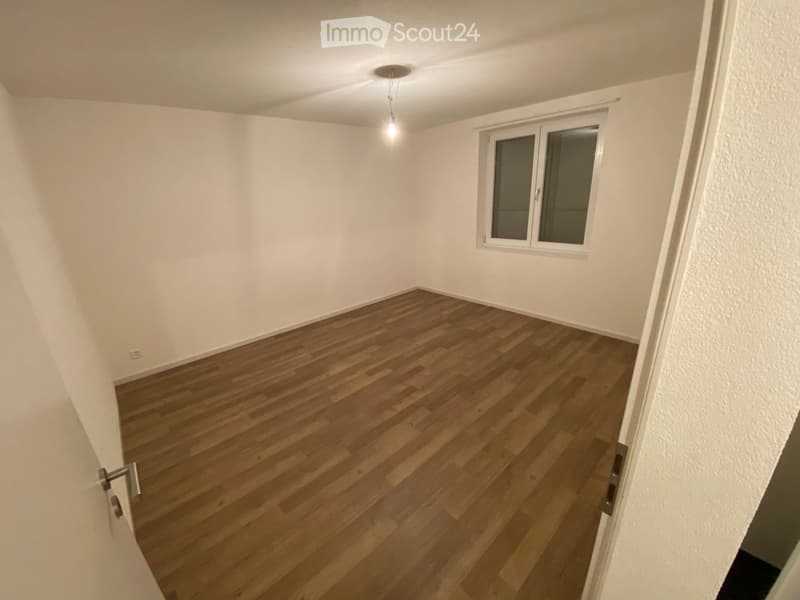 Empty room with wooden flooring, white walls, and a single window with blinds. The room has a ceiling light fixture and a visible electrical outlet on the wall.