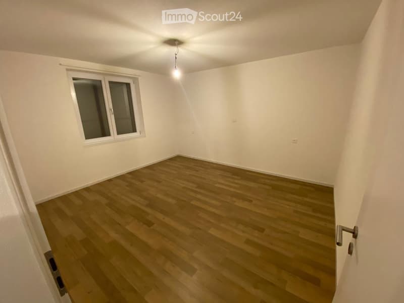 Empty room with wooden flooring, white walls, and a single window with blinds. A ceiling light fixture is visible.