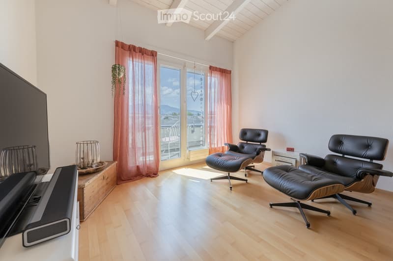 Bright living room with wooden flooring, large windows offering a scenic view, two black leather chairs, a small table, and a television.