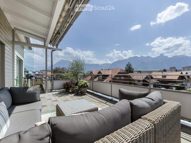 Balcony with a view of mountains, featuring a grey sofa, wicker chair, small table, and several potted plants.