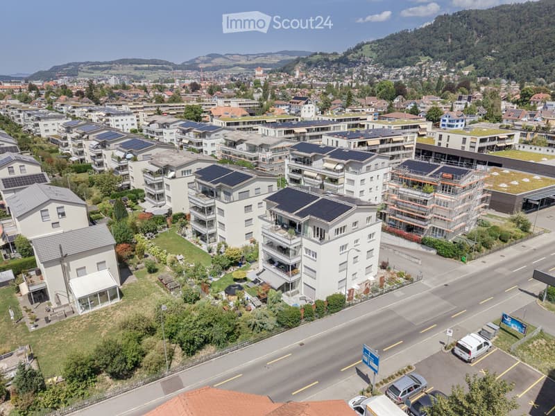 Aerial view of a residential area with multiple apartment buildings, solar panels on rooftops, green spaces, and a road with parked cars.