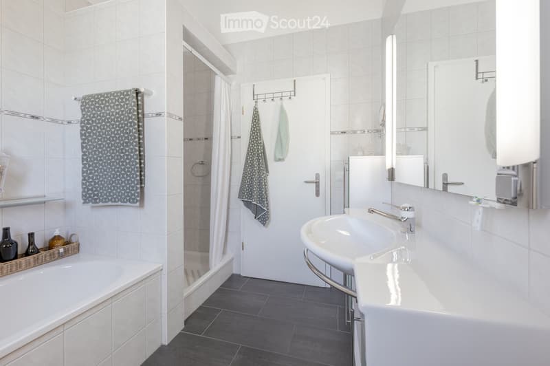 Modern bathroom with white tiles, bathtub, sink, mirror, and towel rack. Features a shower area with a curtain and a small basket with decorative items on the edge of the bathtub.