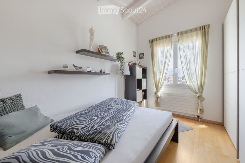 A modern bedroom with a large bed, patterned bedding, and pillows. The room features wooden flooring, white walls, and a large window with light-colored curtains. A black shelf holds decorative items, and a small black cabinet is visible near the window.