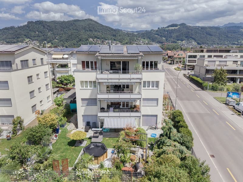 A multi-story house with balconies, surrounded by greenery and a scenic mountain view in the background.