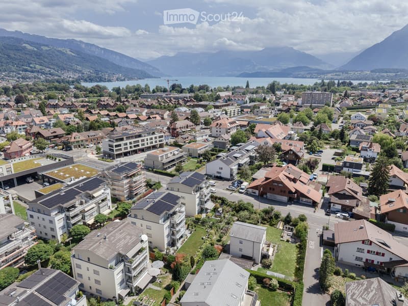 Aerial view of a residential area with houses, apartments, and solar panels on rooftops, surrounded by greenery and mountains in the background.