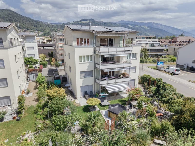 A modern multi-story residential building with balconies, surrounded by greenery and a scenic mountain view in the background.