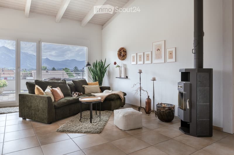 Modern living room with large windows offering a scenic mountain view, a green sectional sofa, a round coffee table, an ottoman, a fireplace, floor lamps, and decorative items including picture frames and a plant.
