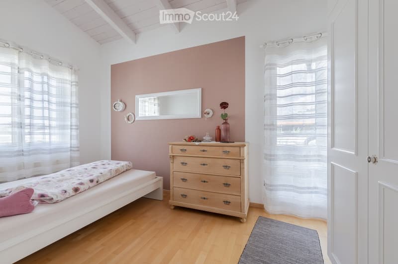 Cozy bedroom with a single bed, wooden nightstand, mirror, decorative vase, and a clock on the wall. Light-colored curtains and a grey rug complete the space.