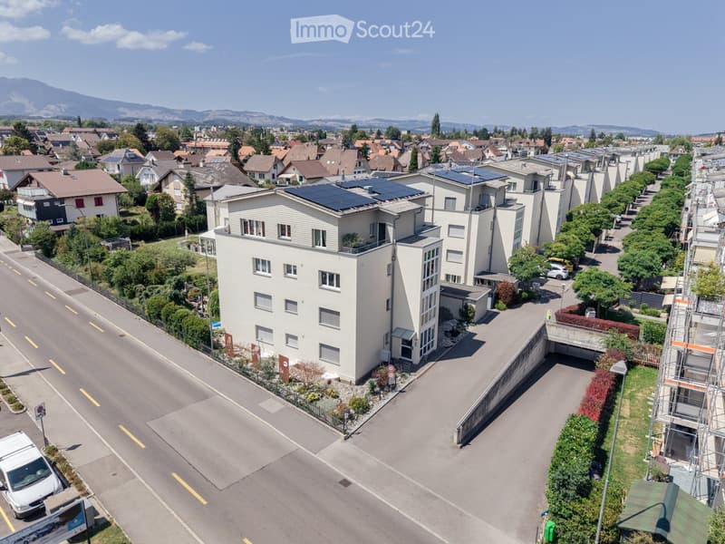 Aerial view of a modern residential building with solar panels on the roof, surrounded by a well-maintained garden and a street with parked cars.