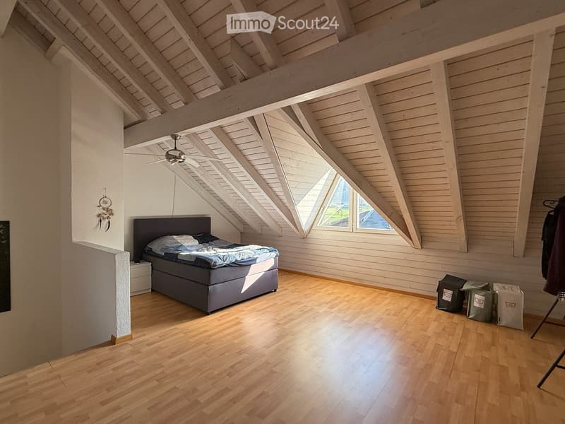 Bright, airy attic bedroom with wooden floors, exposed wooden beams, and large skylight windows. Features a double bed with blue bedding, a nightstand, and a ceiling fan.