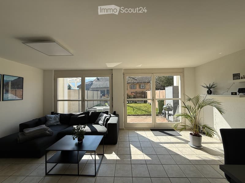 Modern living room with large windows and sliding doors leading to a patio, featuring a black sectional sofa, a black coffee table, and potted plants.