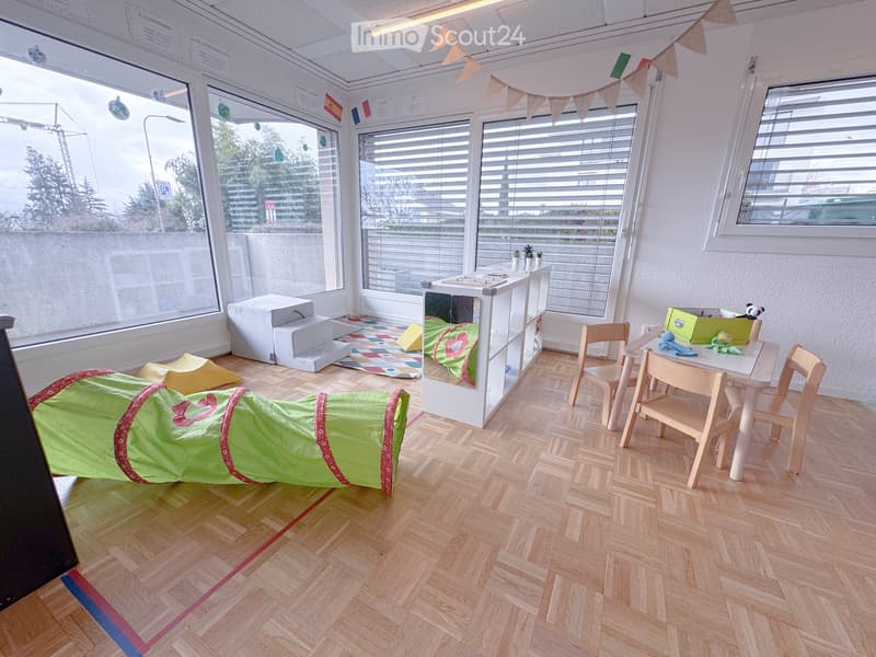 Bright, modern room with wooden flooring, large windows, colorful play area, sofa, small table with chairs, and shelves.