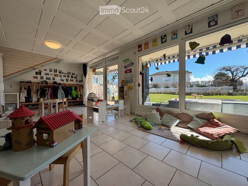 Bright, airy room with tiled floor, large windows, and a cozy seating area with pillows and a toy house on a small table. A coat rack and a small table with chairs are visible, along with a TV screen and a view of a green outdoor area.