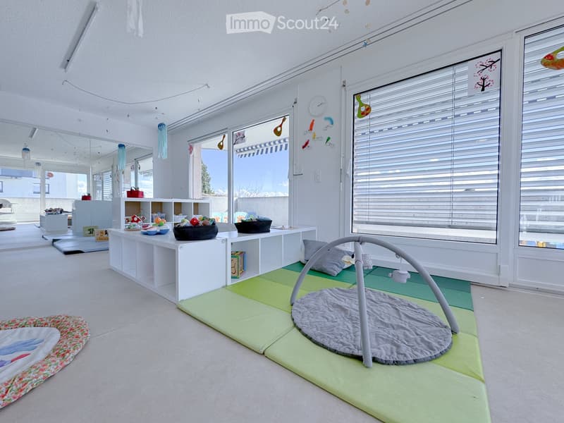 Bright, airy room with large windows, white walls, and a baby gym in the center. Shelves hold toys and storage items, and colorful play mats cover the floor.