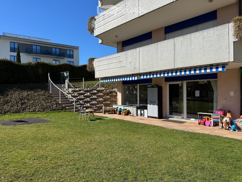 A well-maintained garden area with a grassy lawn, a small patio with a table and chairs, and various children's toys scattered around. The area is adjacent to a modern building with a staircase leading up to it.