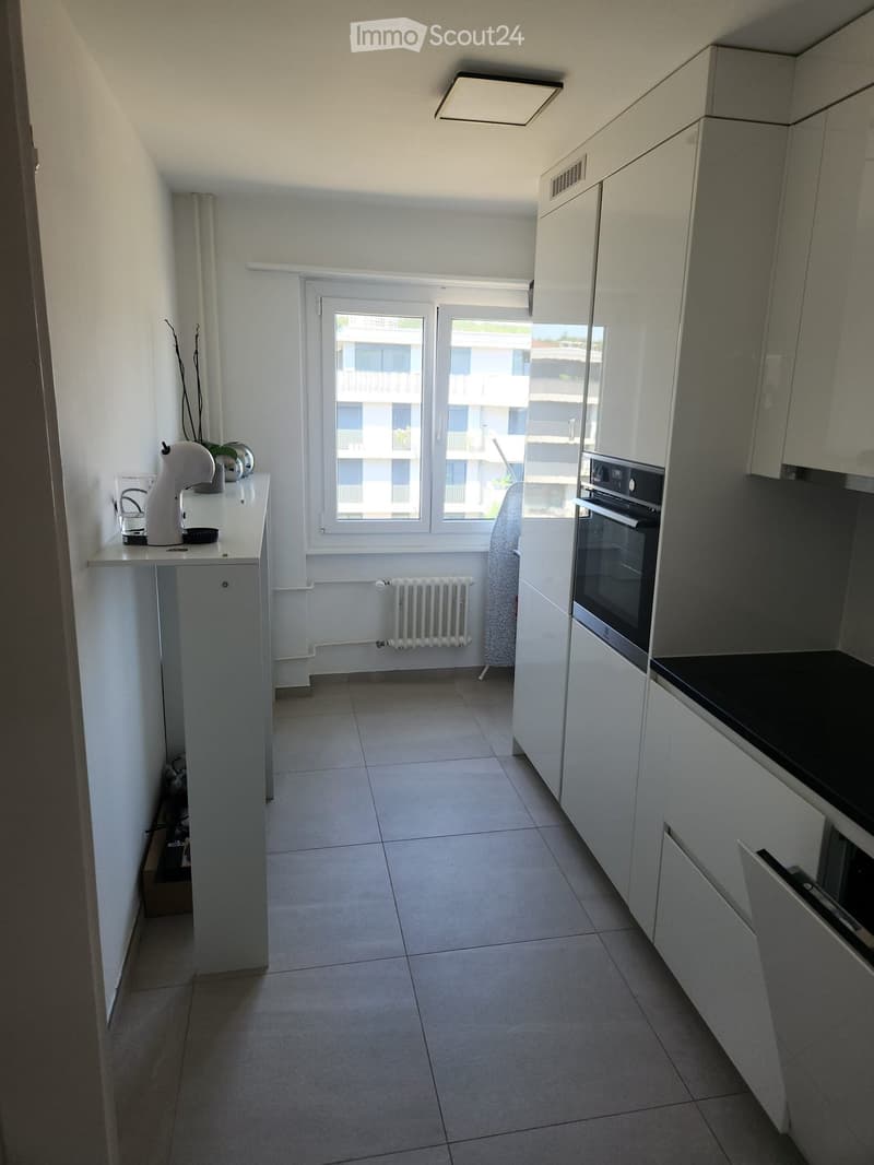 Modern kitchen with white cabinets, tiled floor, and a window overlooking a building. Includes an oven, a kitchen island with a coffee maker, and a cup on the counter.