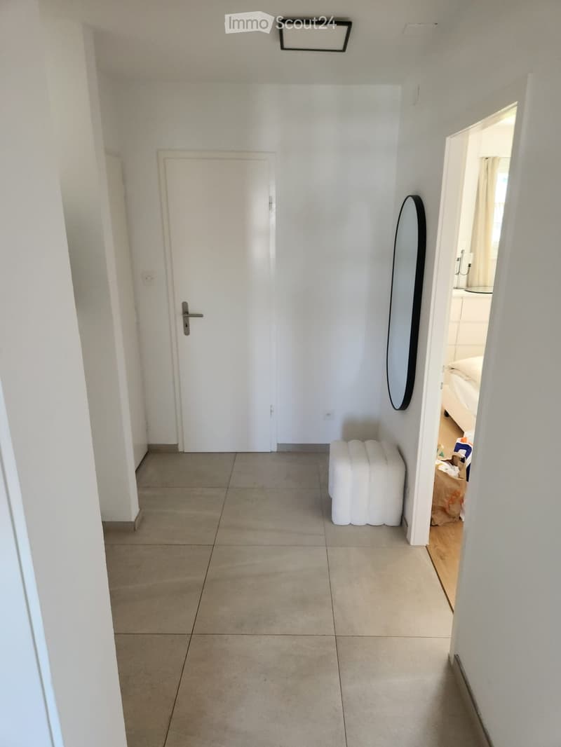 A clean, modern hallway with tiled flooring, white walls, and a closed door on the left. A mirror is mounted on the wall to the right, and a small white bench sits near the doorway leading to another room.