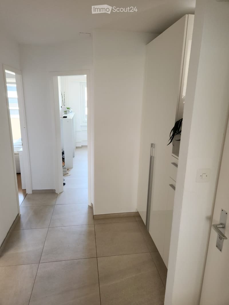 A clean, white-walled hallway with tiled flooring, leading to an open doorway that reveals a glimpse of a furnished room beyond.