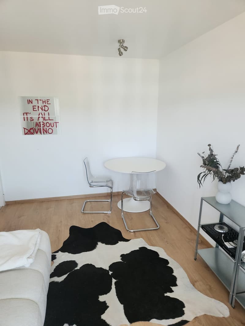 Modern living room with white walls, wooden floor, cowhide rug, sofa, dining table with chairs, shelf with plant and electronic device, and artwork on wall.