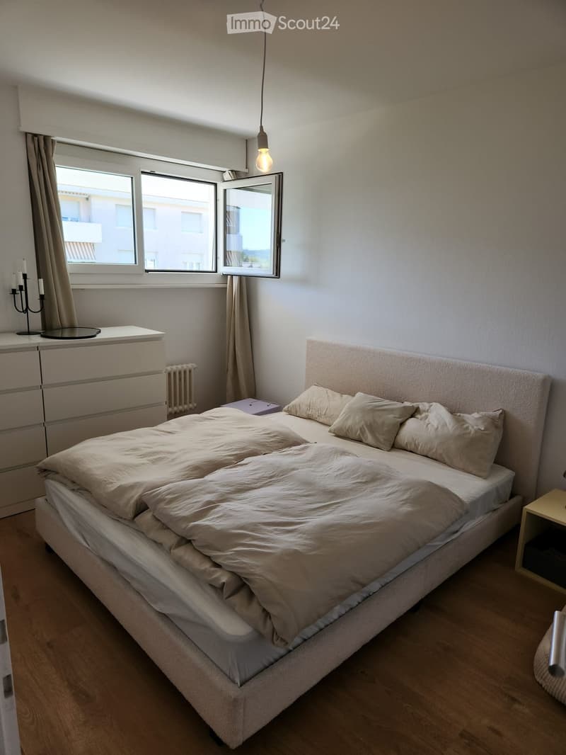 Modern bedroom with a large bed, nightstand, candle holder, and a mirror on the wall. The room features wooden flooring and large windows with curtains.