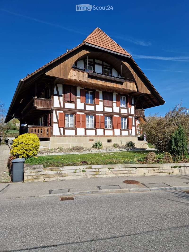 Traditional two-story house with wooden beams, red shutters, and a sloped roof. Features a balcony on the upper level and a well-maintained garden in front.