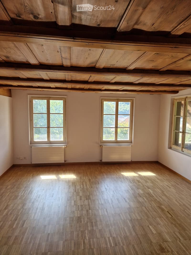 Empty room with wooden floor, exposed wooden ceiling beams, and three windows with radiators beneath them.