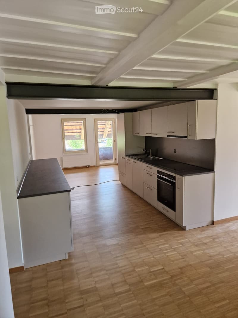 Modern kitchen with light-colored cabinets, a sink, an oven, and a wooden floor. The space is open and well-lit with large windows and a door leading to an outdoor area.