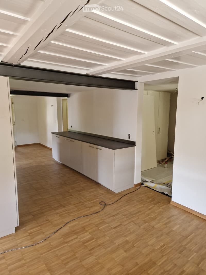 Empty room with wooden flooring, white walls, and a small white cabinet with a black countertop. Exposed beams on the ceiling and visible construction materials in the background.