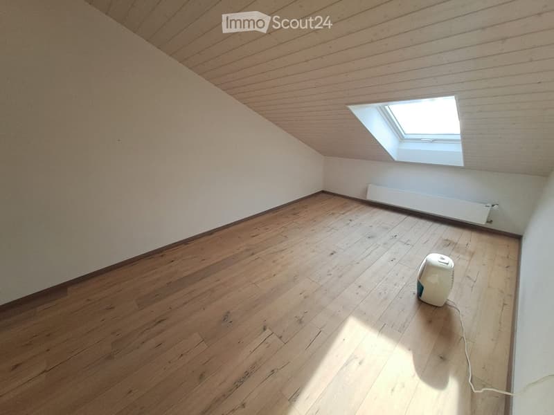 An attic room with wooden flooring, slanted wooden ceiling, and a skylight window. A small white fan is placed on the floor.