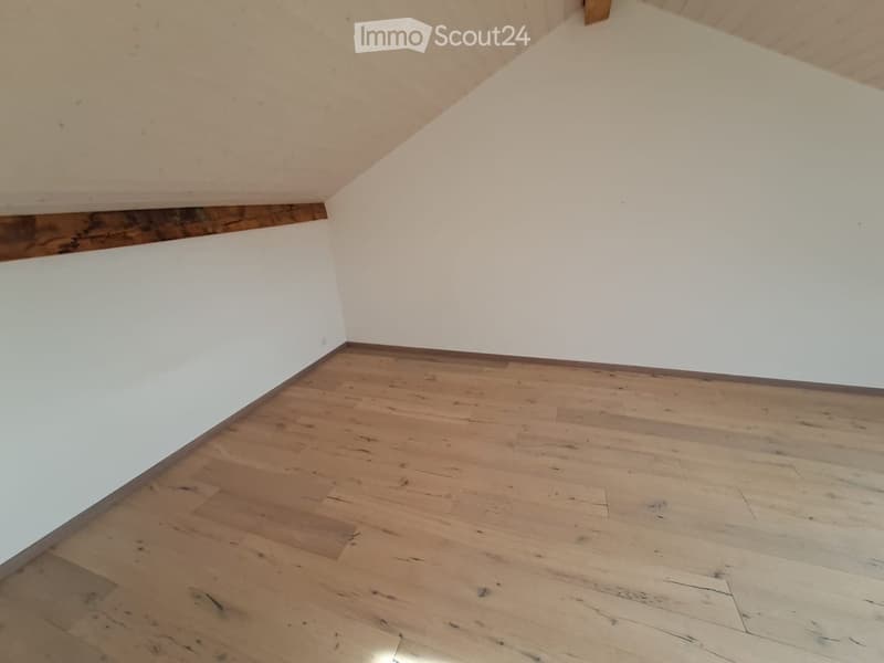 Empty room with wooden flooring and white walls, featuring a slanted ceiling and exposed wooden beams.