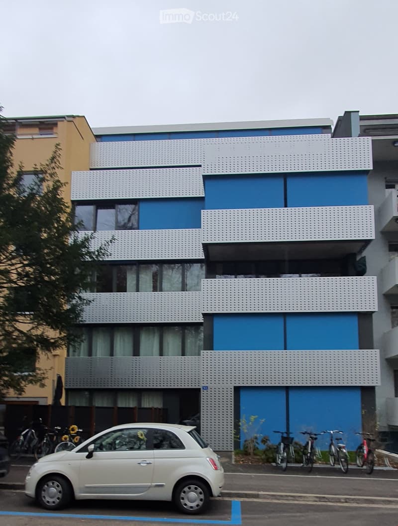Modern multi-story building with blue and white facade, large windows, and balconies. Bicycles are parked in front, and a small white car is parked on the street.