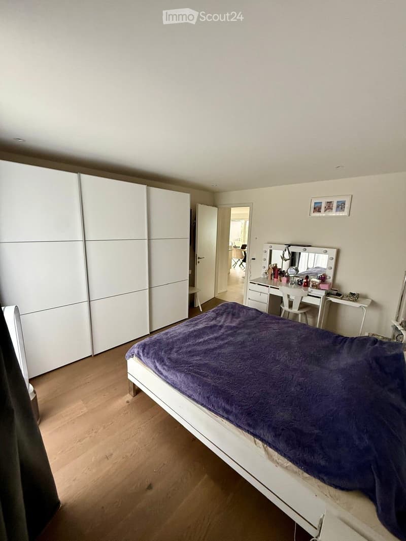 Bedroom with a white bed covered in a purple blanket, a large white wardrobe, a vanity with a mirror, and a desk with a chair.