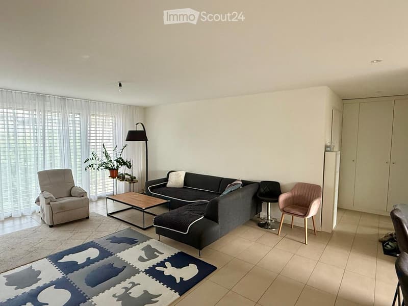 Modern living room with a grey sectional sofa, coffee table, armchair, stool, and a floor lamp. The space features large windows with white curtains and a colorful rug on the tiled floor.