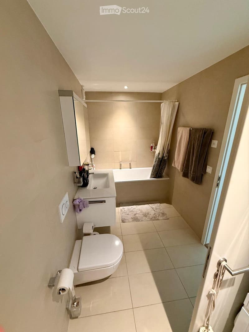 Modern bathroom with a toilet, sink, bathtub, mirror, and towels. Features a sleek design with beige tiles and a small window.