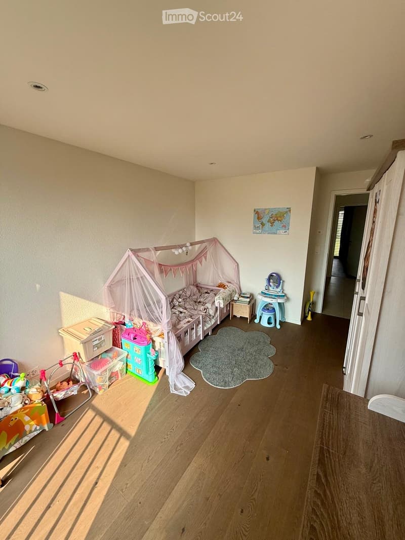 A well-lit children's bedroom with a wooden floor, a bed with a canopy, a nightstand, a small rug, and various toys including a toy castle, a toy crib, and a toy car. A world map is displayed on the wall.