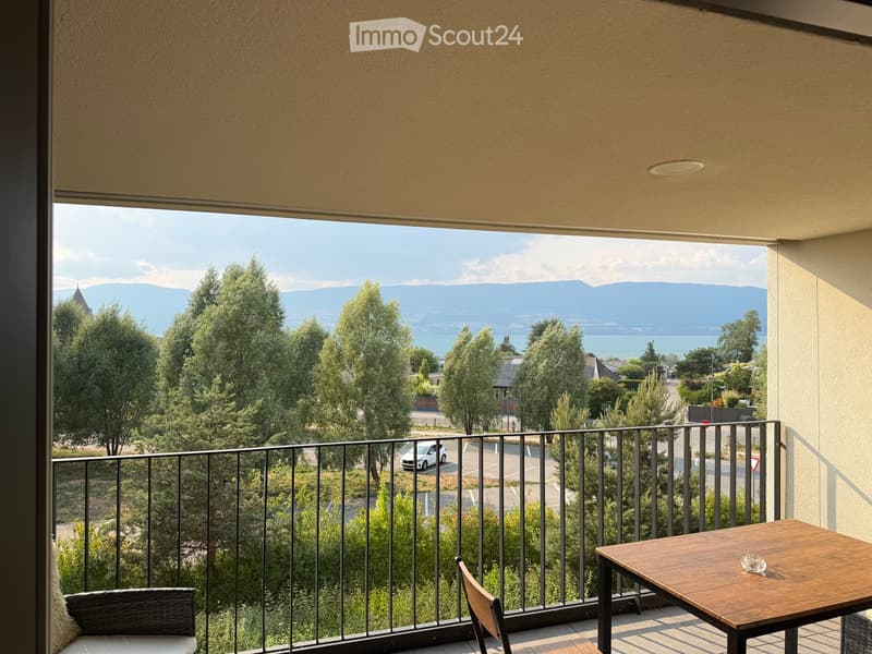 Balcony with wooden table and chairs, overlooking a scenic view of trees, a lake, and distant mountains.