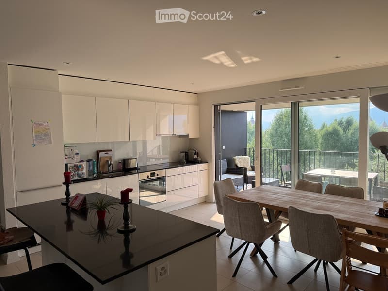 Modern kitchen with white cabinets, black countertop, dining table with chairs, and a view of greenery through large windows.